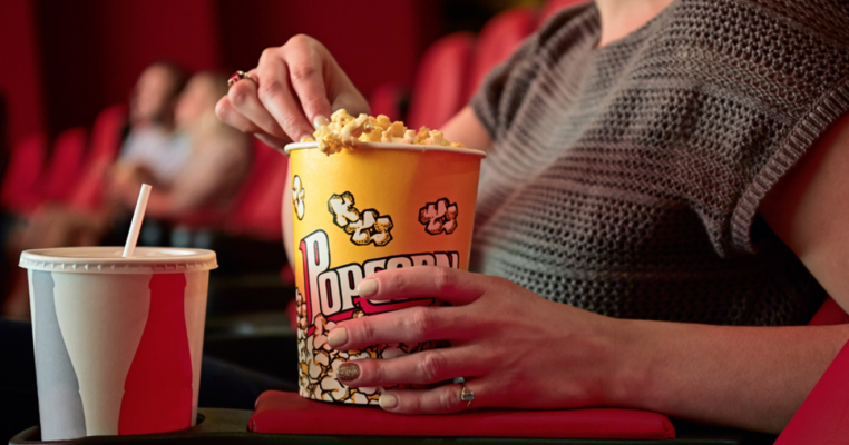 uma pessoa na sala de cinema, segurando um vaso de pipoca escrito "popcorn", com um copo de refrigerante à frente