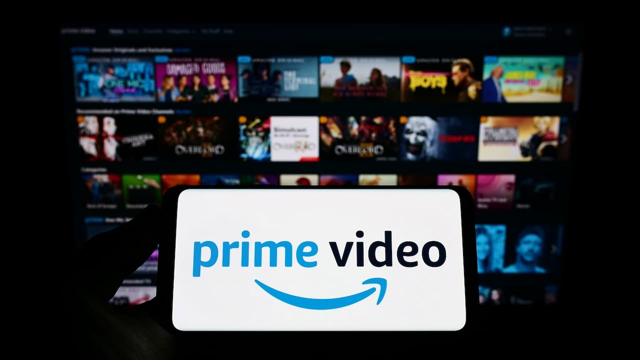Celular mostrando na tela a logo do prime video e ao fundo, desfocado, uma TV com o catálogo do prime video. Amazon Prime Video
