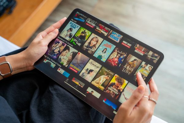 Mãos segurando um tablet com o catálogo de uma plataforma de streaming com filmes e séries.