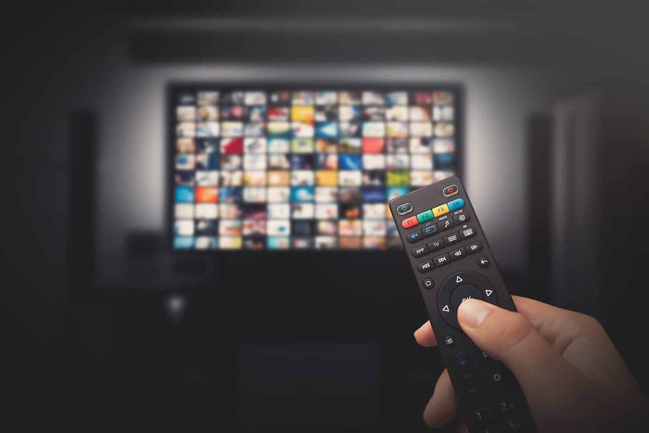 TV ao fundo desfocada, com diversas telas para escolher um filme ou série. Em primeiro plano uma mão segurando um controle remoto.