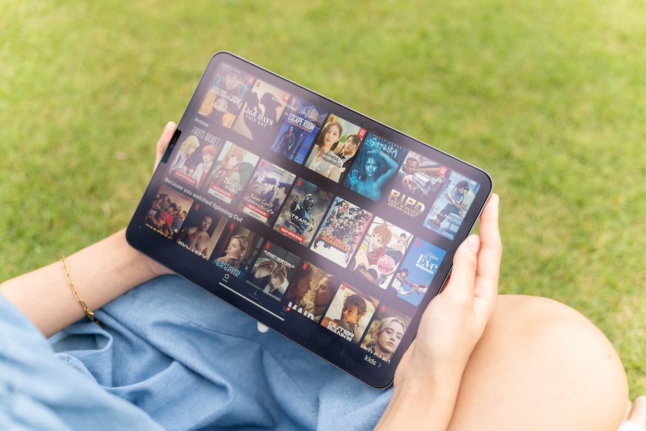 Uma mulher segurando um tablet, aparentemente em um parque. O tablet está com a tela aberta no catálogo com séries, filmes e animes.
