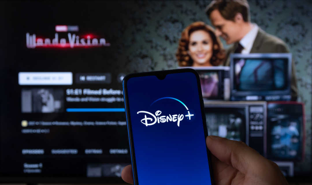 Celular com logo da Disney+ em frente a uma smart TV mostrando a tela inicial do Disney+