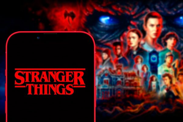 Celular com a logo "Stranger Things" e ao fundo, desfocado, o pôster da série, com os atores principais e vilão.
