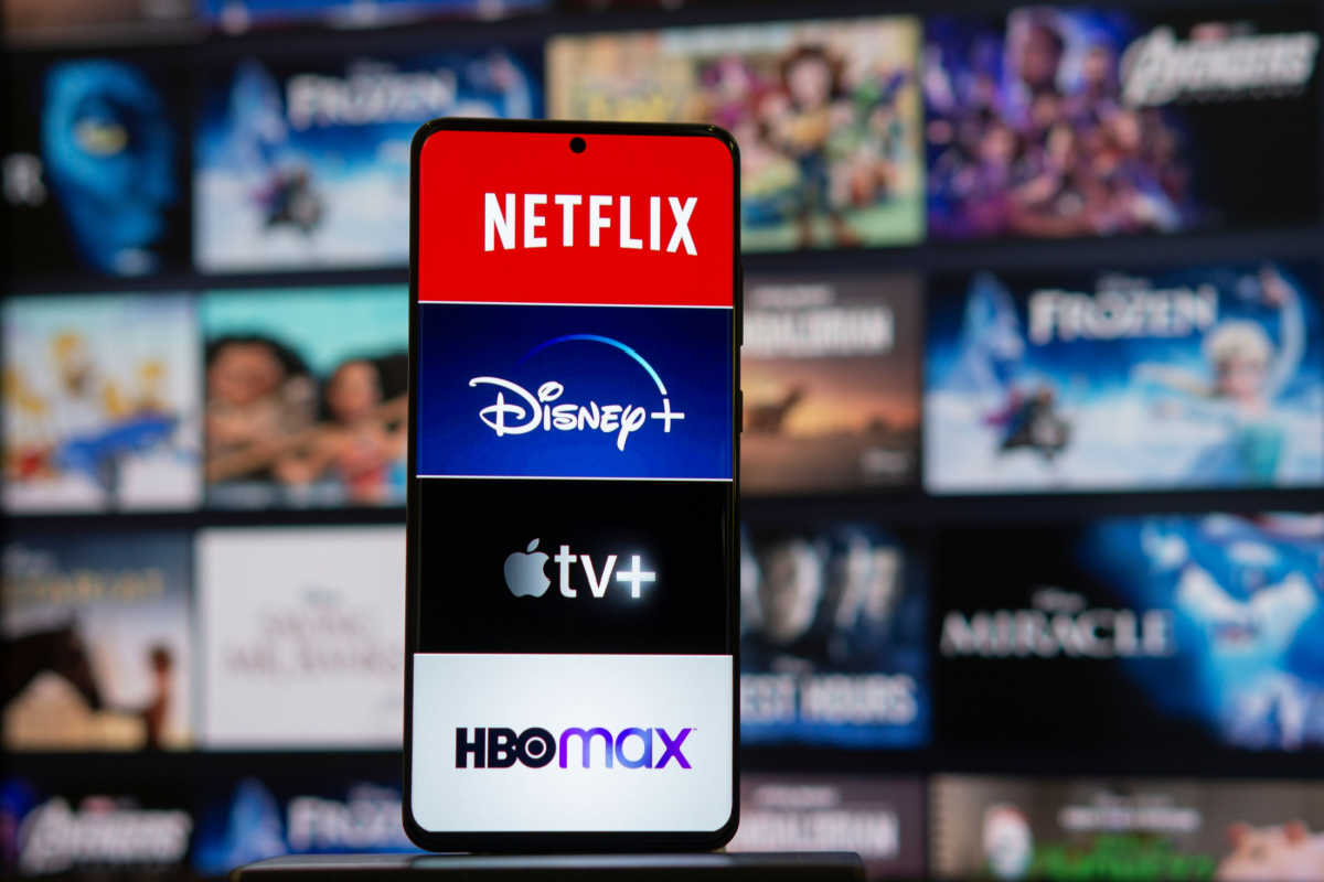 Imagem de um celular com logos da Netflix, Disney+, Apple TV+ e HBO Max e, ao fundo, uma tela com várias capas de produções visuais