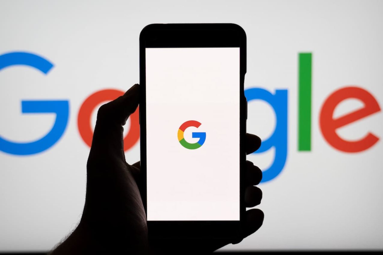 silhueta de mão segurando um smartphone com o logo do Google. Em segundo plano um telão escrito Google.