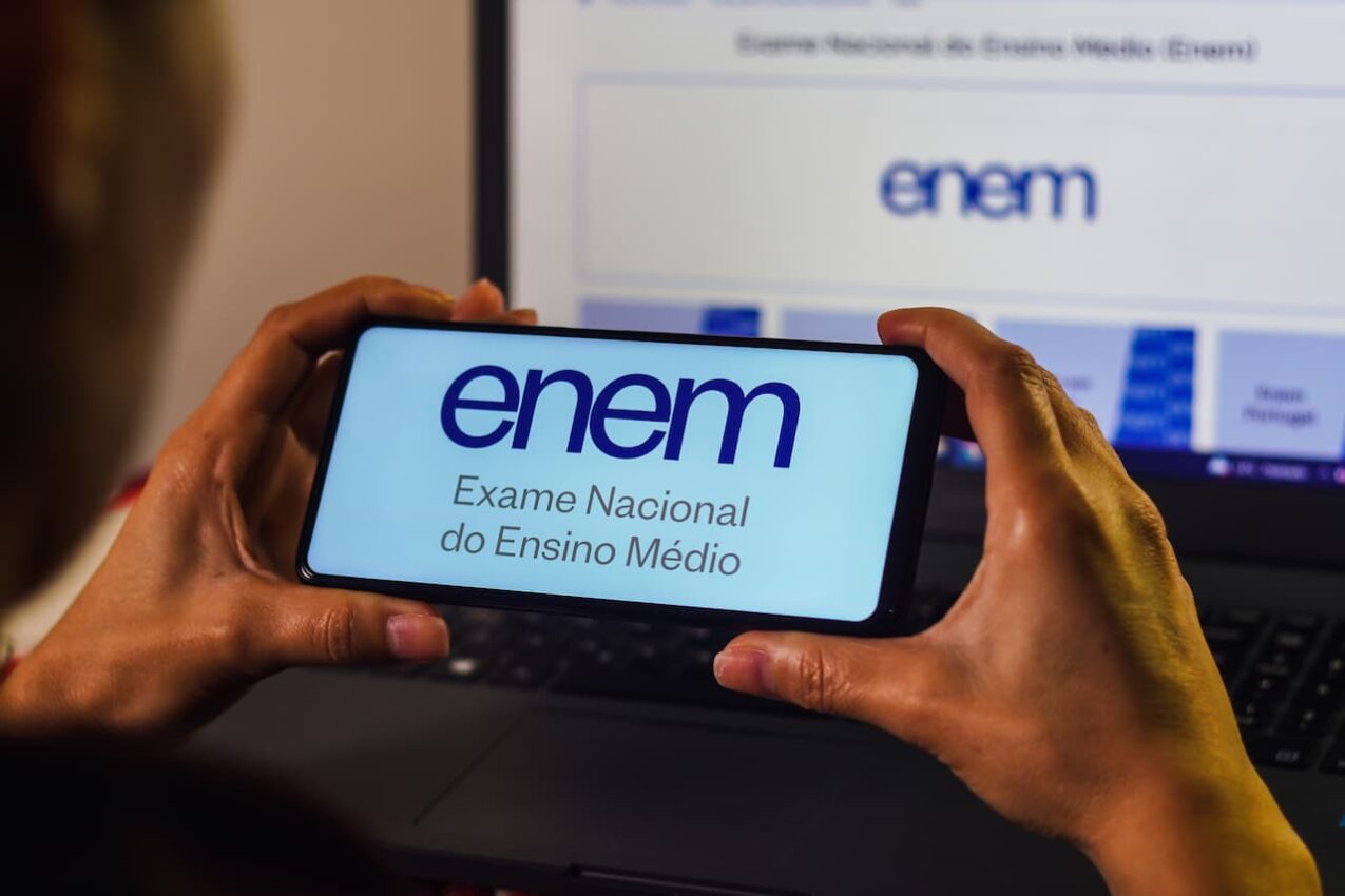 Mãos segurando um celular na horizontal, na tela está escrito ENEM. Ao fundo, uma tela de computador com o site do Enem aberto.