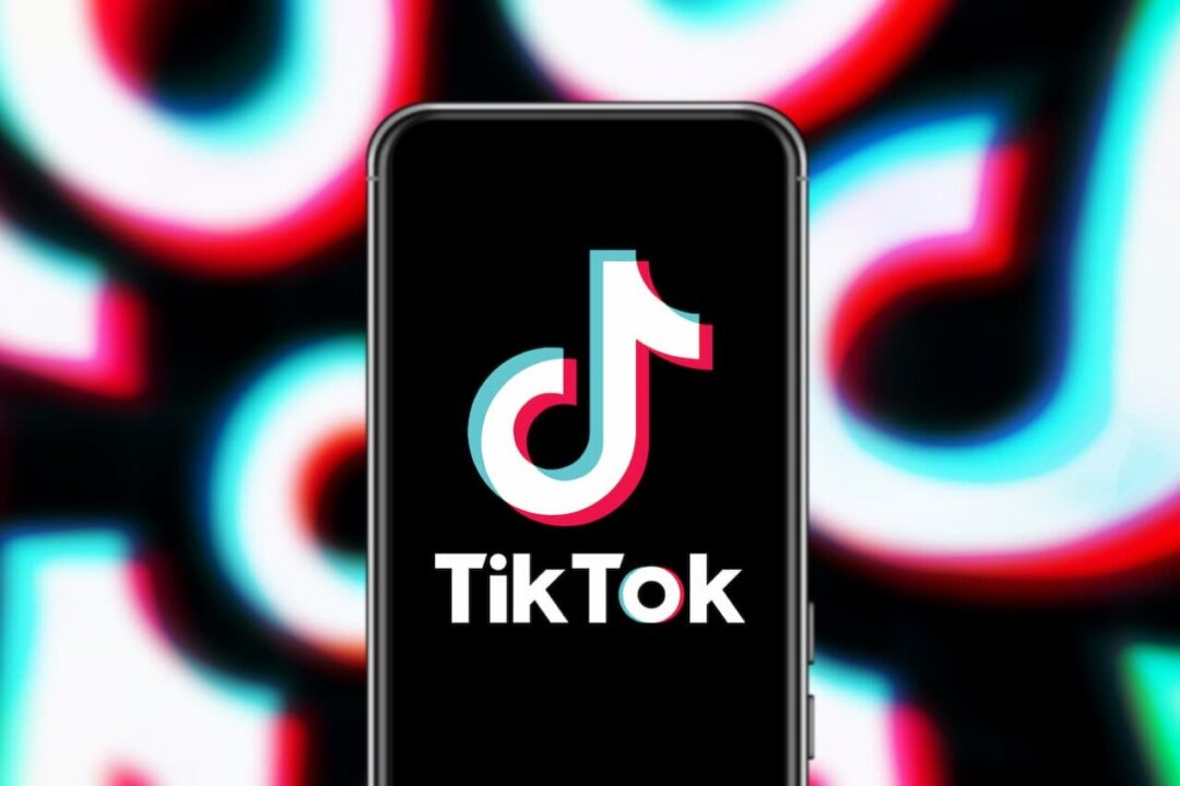 Aplicativo TikTok aberto no celular, com a logo a amostra. Ao fundo, símbolos do app desfocado.